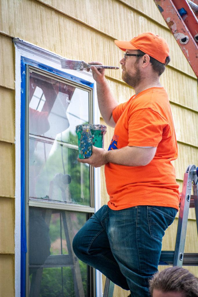 A volunteer on a ladder in an orange t-shirt and hat holds a paint brush and bucket, painting window trim in white. The home has yellow siding.