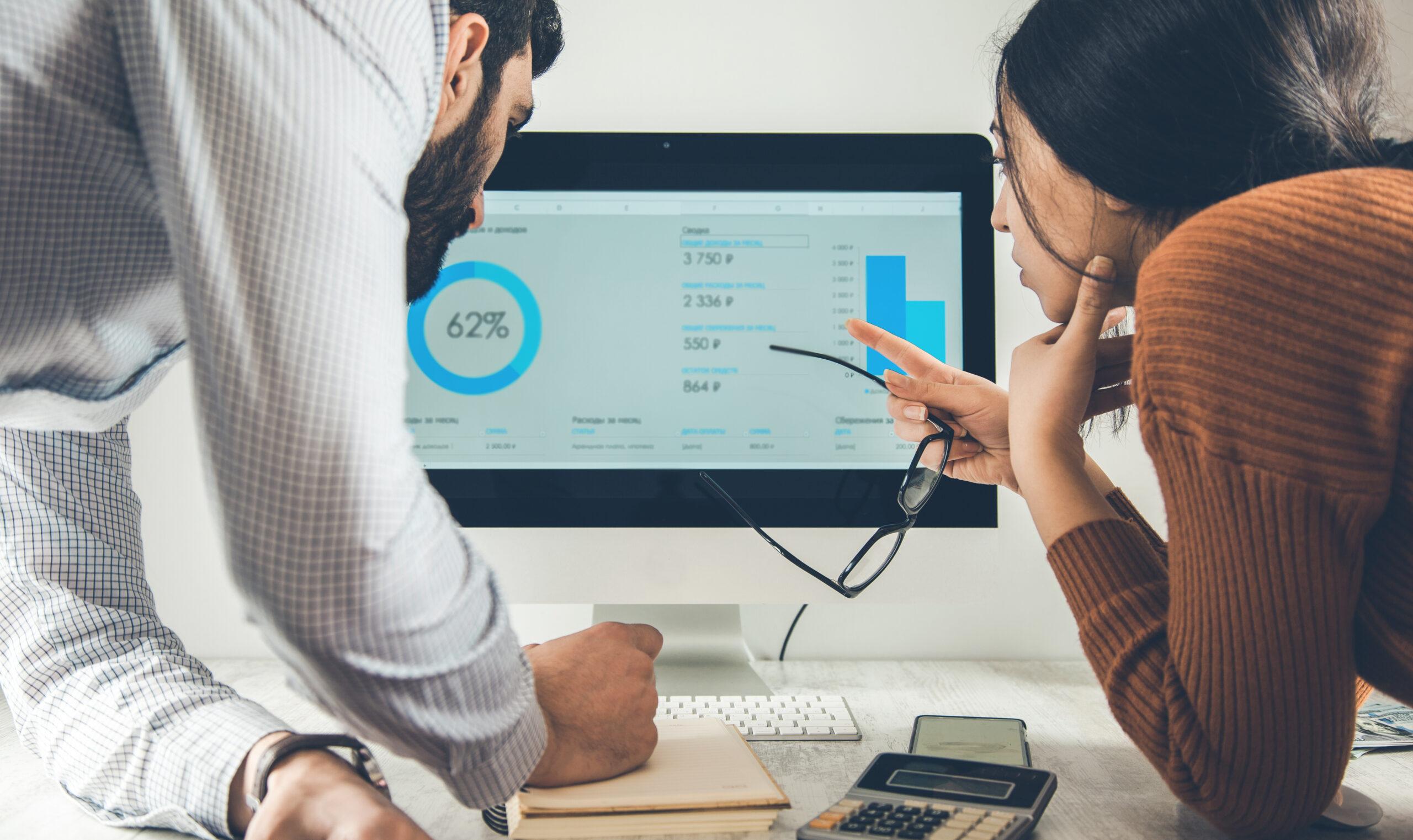 Close up of 2 people looking at data on a computer screen. She is on the rigth holding glasses and pointing to the data. They are leaning on a desk.