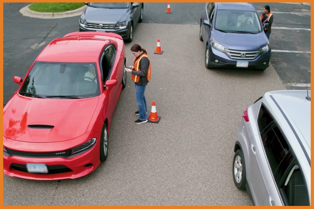 cars lined up for drive thru voting. A person checks a red car in.