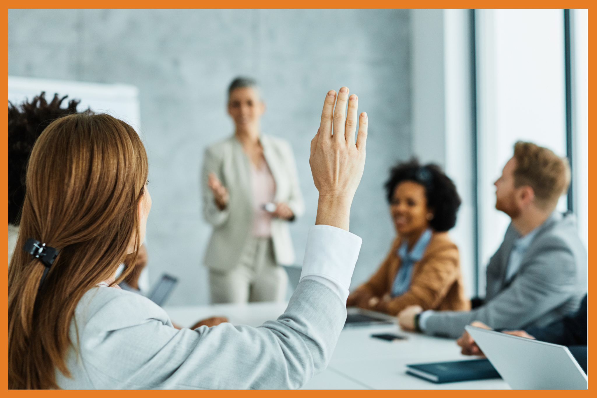 A person raises their hand to second a motion during a business meeting while a group of colleagues listens to a speaker at the front of the room. Everyone is wearing business attire and appears engaged.